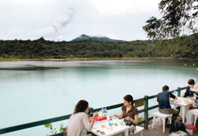 火山から噴き出る白煙を背景に、天気によって３色に見えるリノウ湖畔で食事をする観光客＝ミナハサ県で