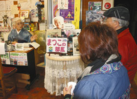 矢島駅の一角の売店で佐藤まつ子さん（左）の話に耳を傾ける観光客＝いずれも秋田県由利本荘市で