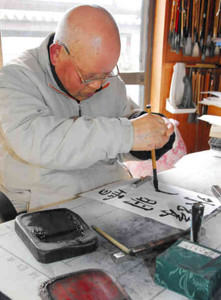 利き手と逆の左手で筆を握り、右手を添えながら字をしたためる田沢豊和さん＝可児市緑ケ丘で