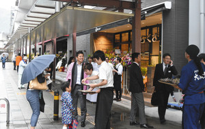 開業を迎え、店舗前で通行人に試食品を配るスタッフら＝大津市のＪＲ大津京駅前で