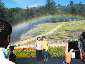 御嶽山を背景に散水して作る虹と利用客を撮影するサービス「いってらっしゃいレインボー」＝４日、木曽町の御岳ロープウェイで（中央の被撮影者はスタッフ）