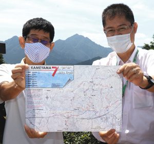 完成した登山地図を持つ松永さん（左）ら。後ろに見えるのが「亀山七座」の一つの錫杖ケ岳＝亀山市関町の観音山公園で