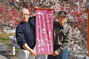 鮮やかに咲くハナモモの前で、手ぬぐいを広げる熊谷代表（左）ら＝阿智村で