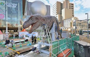 ビルが立ち路面電車が走る中で設置作業が始まったティラノサウルスの恐竜ロボット＝福井市のＪＲ福井駅西口で